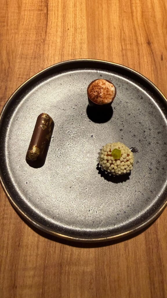 Three small bites arranged on a dark round plate at Evvai in São Paulo.