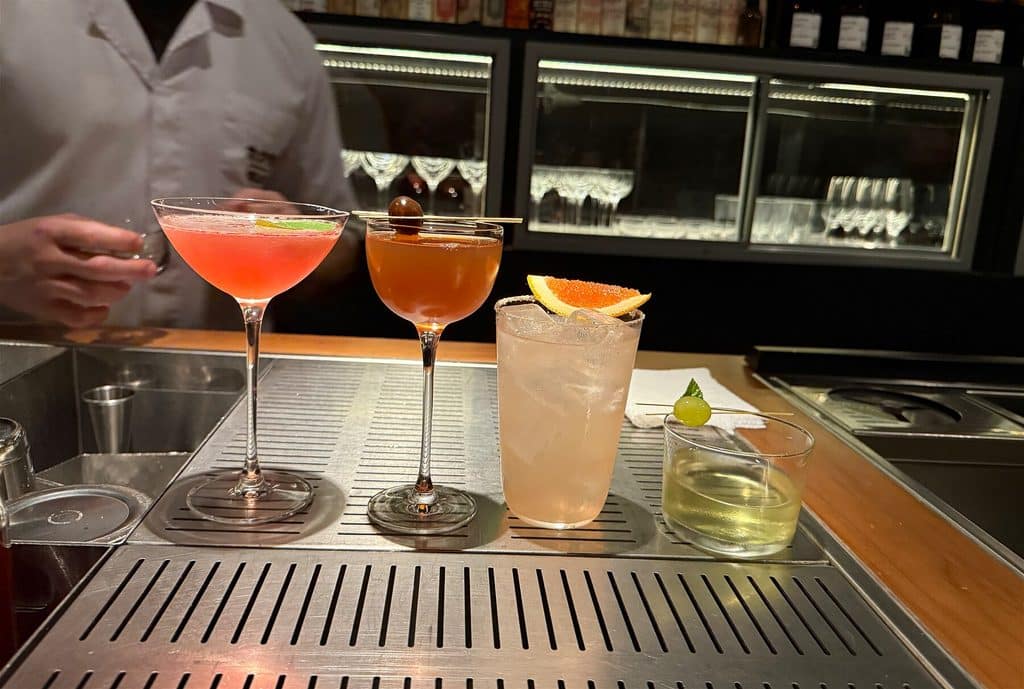 Four colorful cocktails lined up on the bar at Tan Tan São Paulo, with bottles and glassware behind them.
