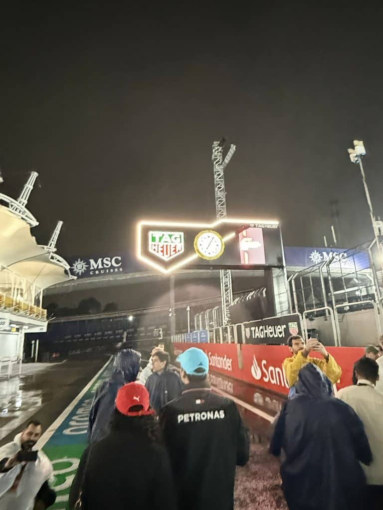 History of Autodromo Jose Carlos Pace Fans walking along the wet Interlagos pit lane beneath an illuminated TAG Heuer clock at night.