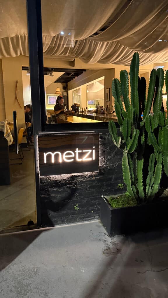Illuminated Metzi entrance sign beside tall cacti outside the restaurant at night.
