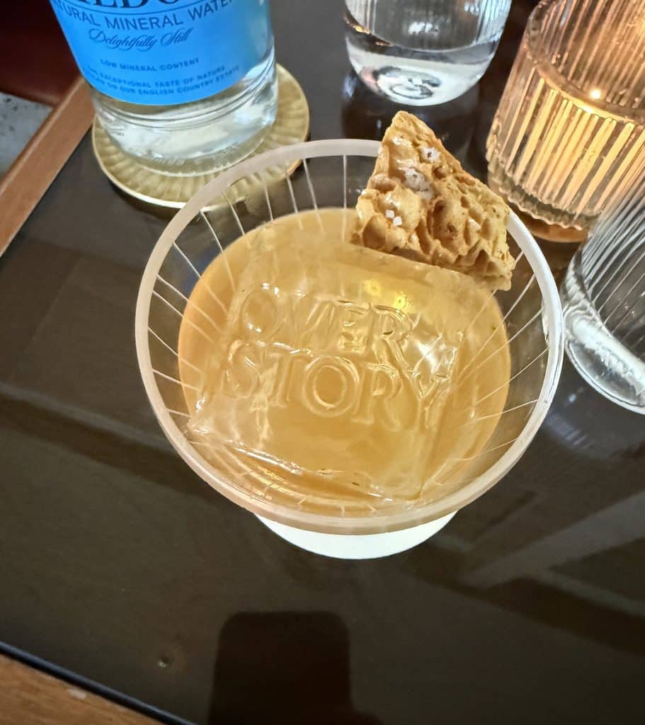 A golden cocktail in a glass marked Overstory beside a candle on a dark table.