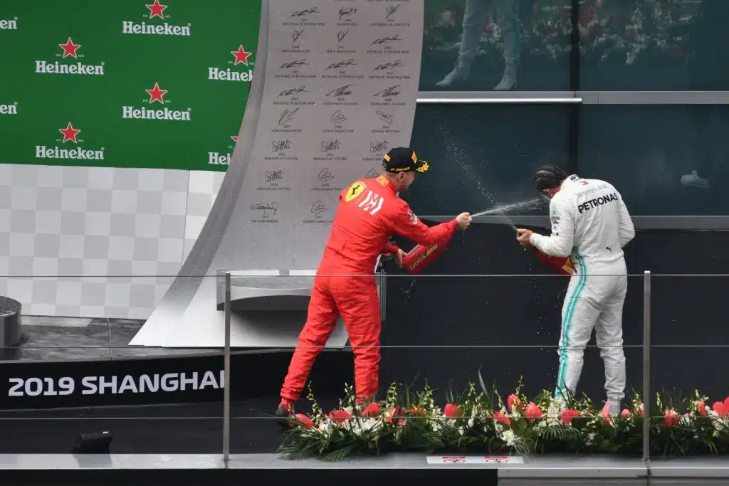 Lewis Hamilton Paintball Story Sebastian Vettel in Ferrari red sprays champagne toward Lewis Hamilton in white Mercedes overalls on the 2019 Chinese