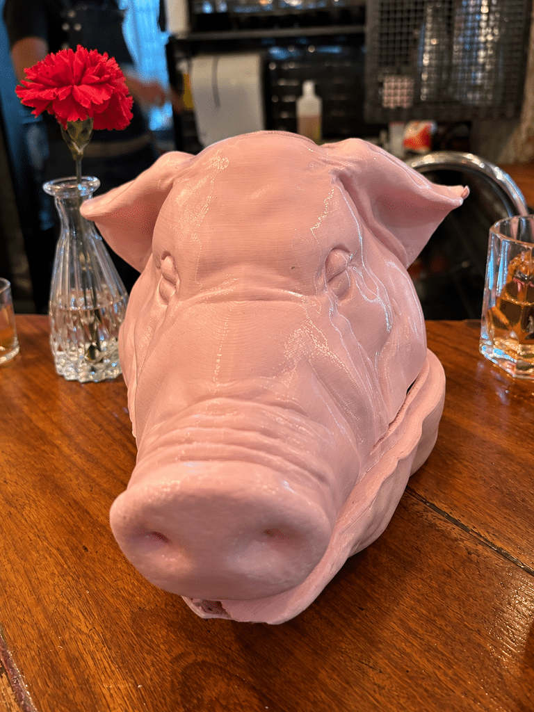 A Casa do Porco review: Closed pink pig’s-head serving piece on the wooden bar at A Casa do Porco, with a red flower and drinks behind it.