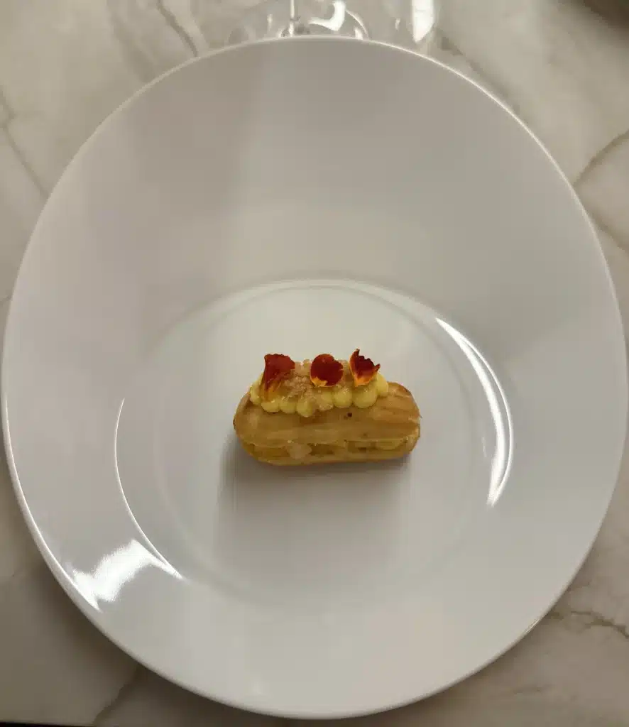 Nelita São Paulo A small golden pastry topped with yellow cream and red-orange petals on an oval white plate at Nelita.