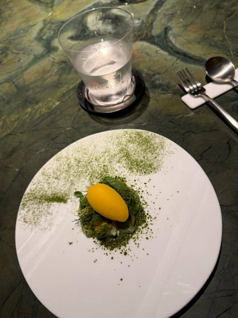 Yellow mango course with green herbs and powder on a white plate at Tuju.