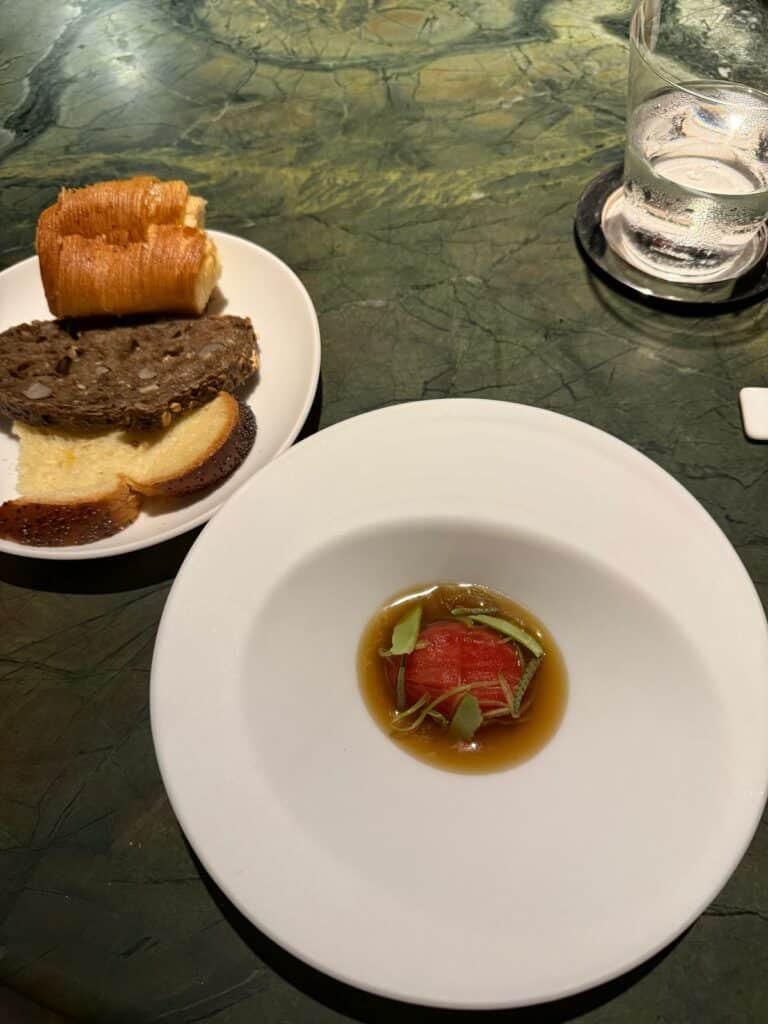 Red tomato course with herbs and broth in a white bowl at Tuju, with bread alongside.