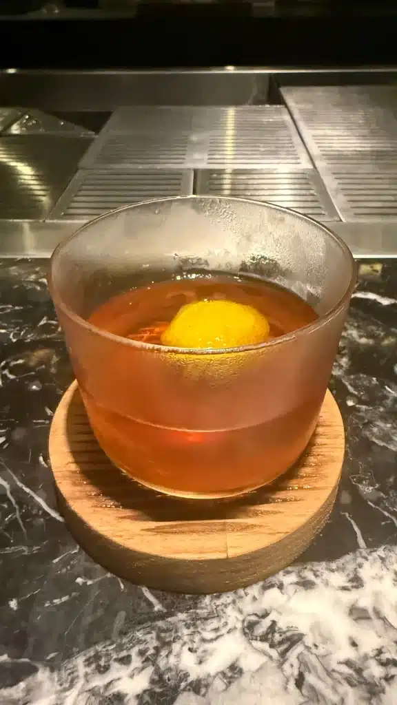 FROM SUPERCARS TO SUITCASES An amber cocktail with a round citrus garnish in a rocks glass on the marble bar at The Liquor Store.