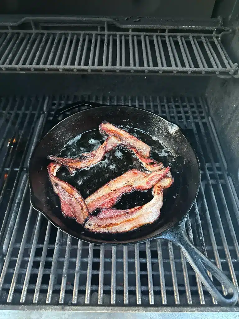 Richard Jarocki Bacon sizzling in a black cast-iron skillet on an outdoor grill.