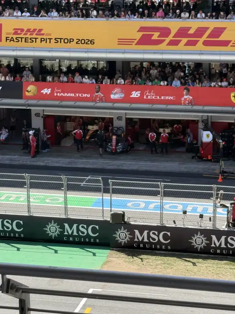 Travel Research Ferrari garages and the pit lane at the Barcelona circuit, viewed from spectator seating behind fencing and a rail.