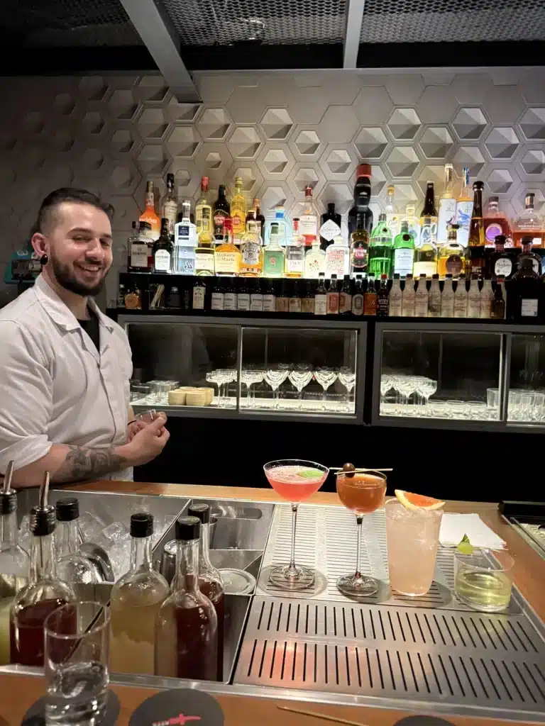 Travel Research A bartender behind the counter at Tan Tan in São Paulo, with bottles, glasses and prepared drinks around him.