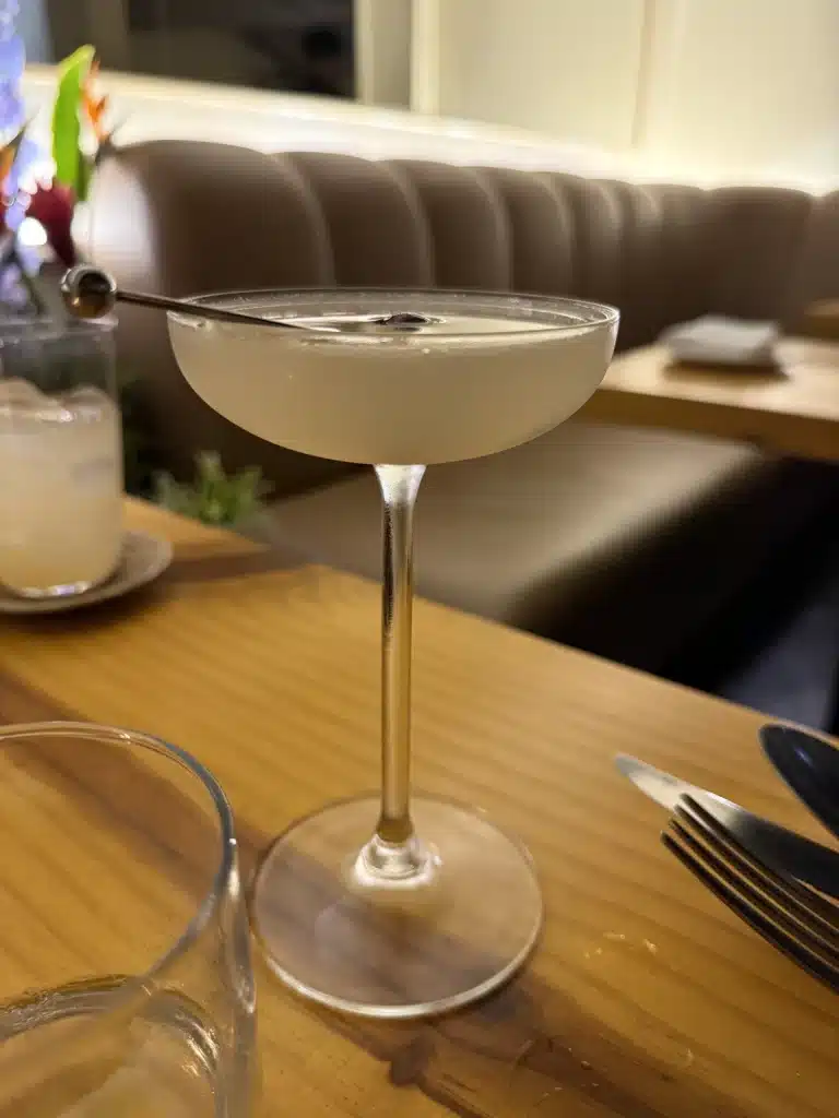 Richard Jarocki A cocktail at Metzi in São PauloA pale cocktail in a coupe glass on a wooden table, with a curved upholstered banquette behind it.