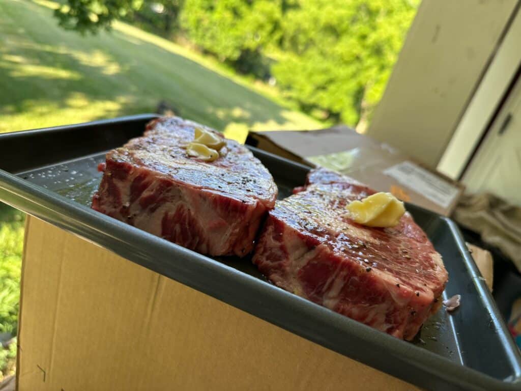 Raw seasoned steaks topped with butter on a tray before grilling