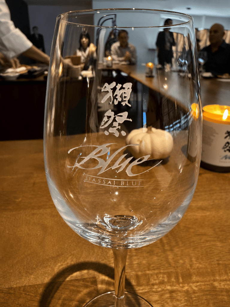 Etched Dassai Blue sake glass at the omakase counter with guests in the background.