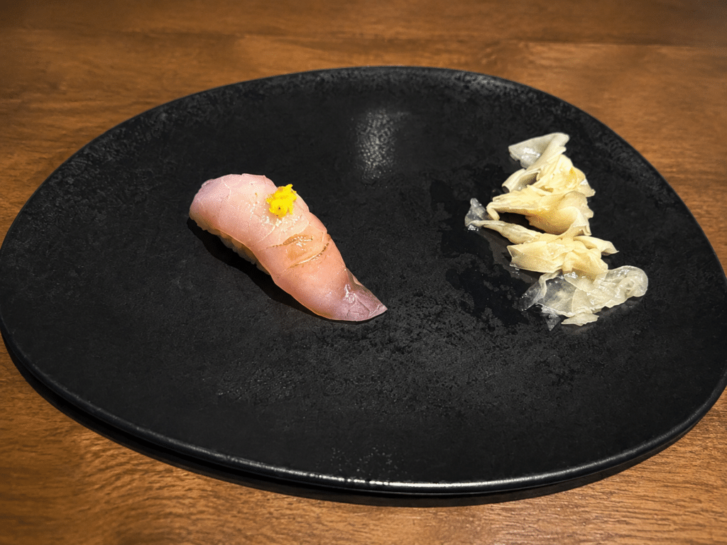 Pale crosshatched nigiri topped with a small garnish and served with pickled ginger on a black plate