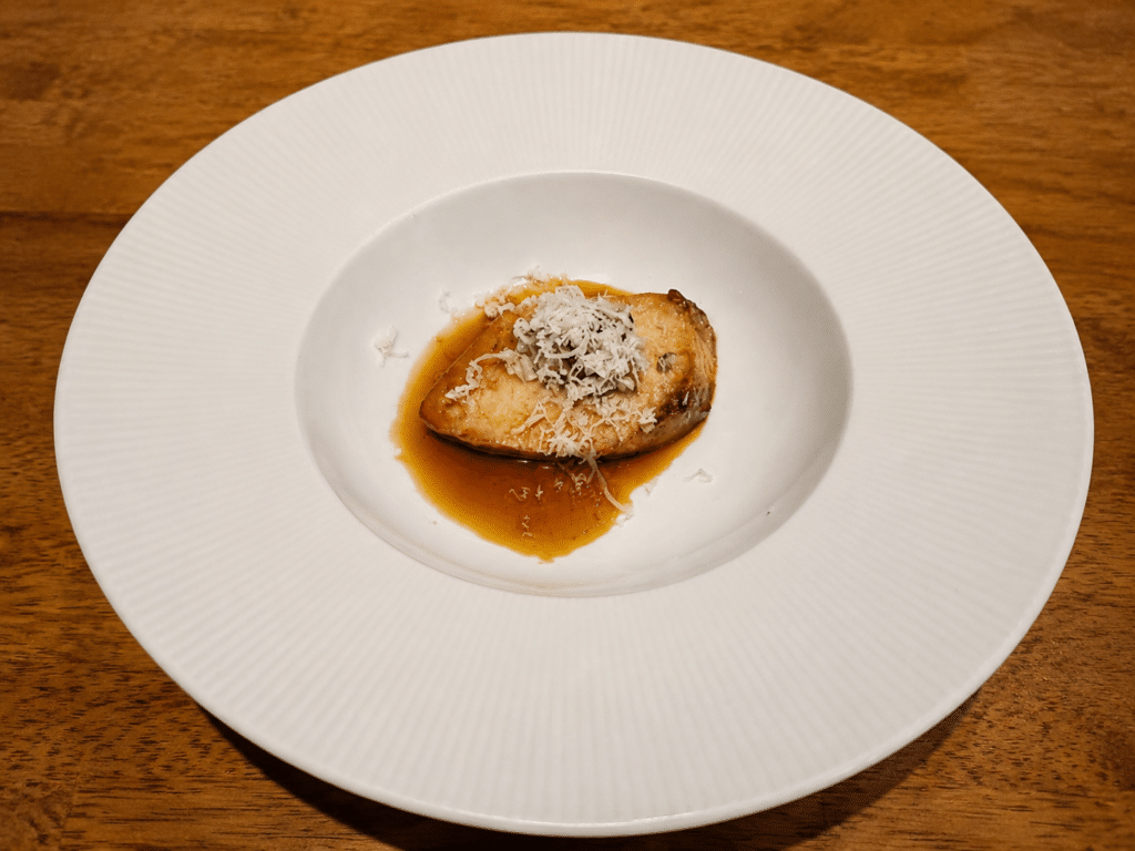 Grilled buri served warm in teriyaki sauce with a finely shredded garnish in a white bowl.