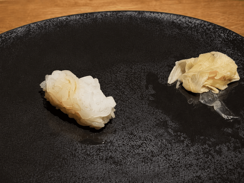 White ika squid nigiri with delicate scoring and pickled ginger on a black plate.