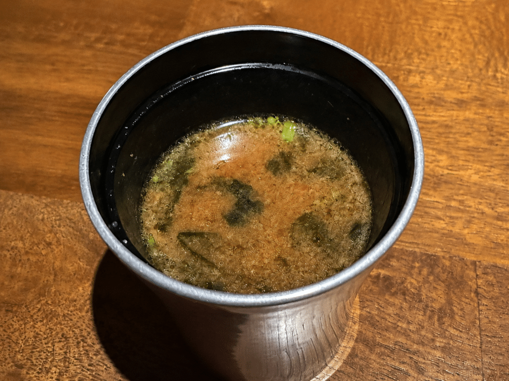 Miso soup served in a metal cup on the wooden omakase counter.