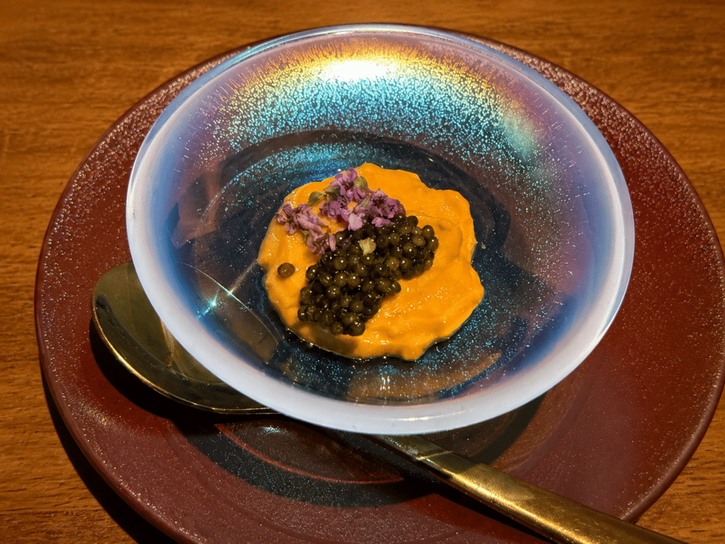 Orange sea urchin cream topped with black caviar and purple edible flowers in an iridescent glass bowl.