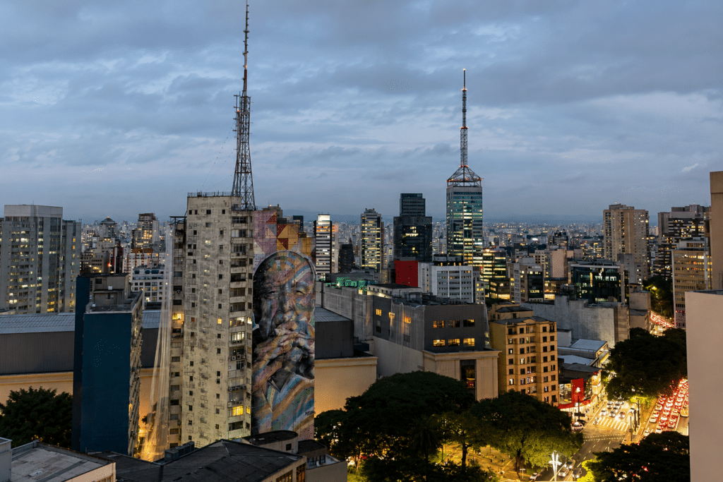 São Paulo skyline at dusk with illuminated buildings, antenna towers and a large mural