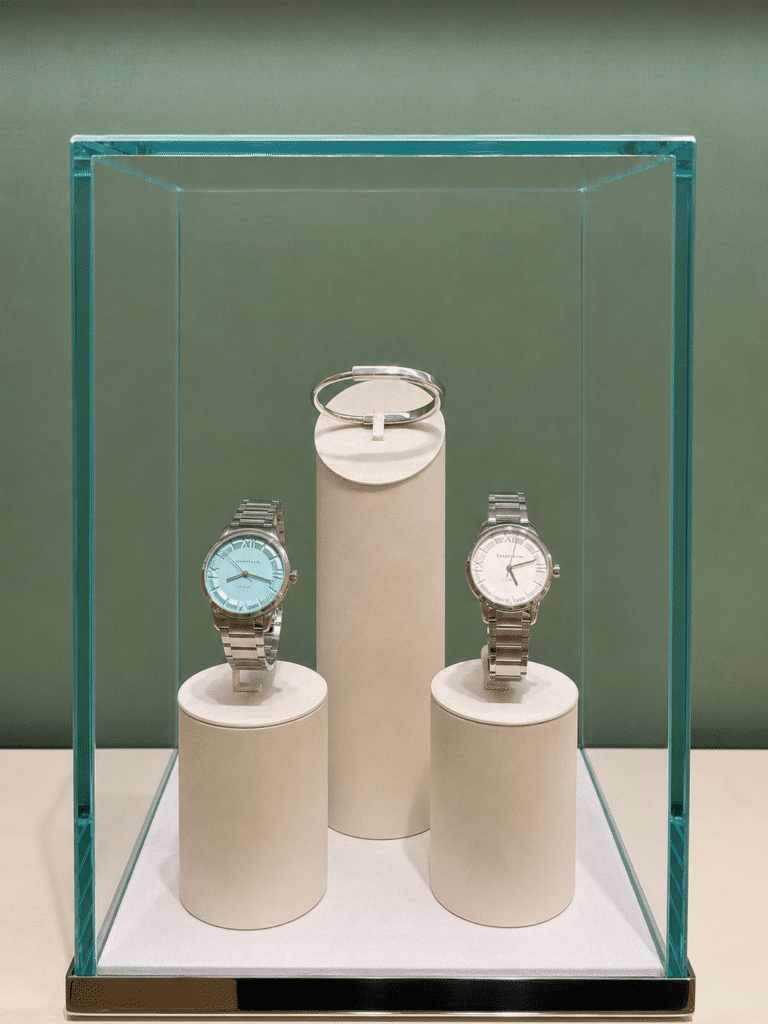 Two stainless-steel Tiffany watches with Tiffany-blue and white dials displayed beneath a silver bracelet inside a glass case