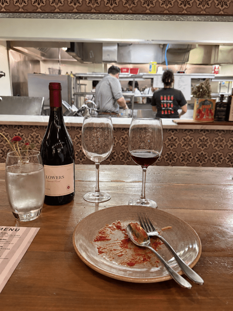 Nearly empty pasta plate with sauce remnants, a fork and spoon, with wine and Viaggio’s open kitchen behind.
