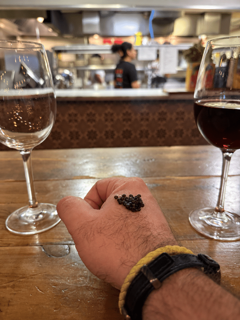 Viaggio x La Valle Dinner A small mound of caviar on the back of my hand between two stemmed glasses, with Viaggio’s kitchen softly blurred behind.