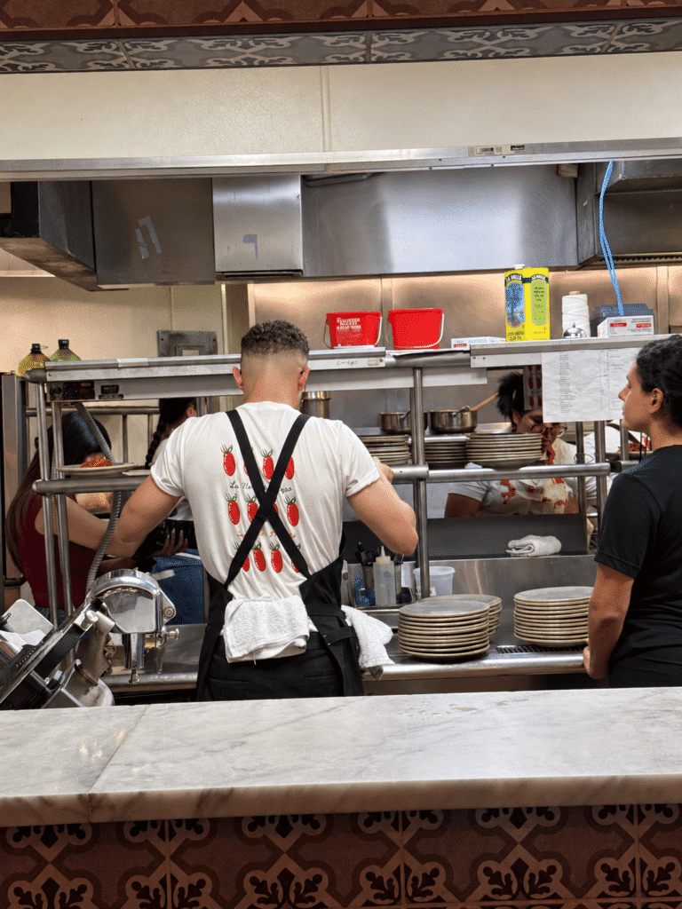 Chef Rabbit seen from behind in a tomato-print shirt and black apron, plating alongside the team in Viaggio’s kitchen.