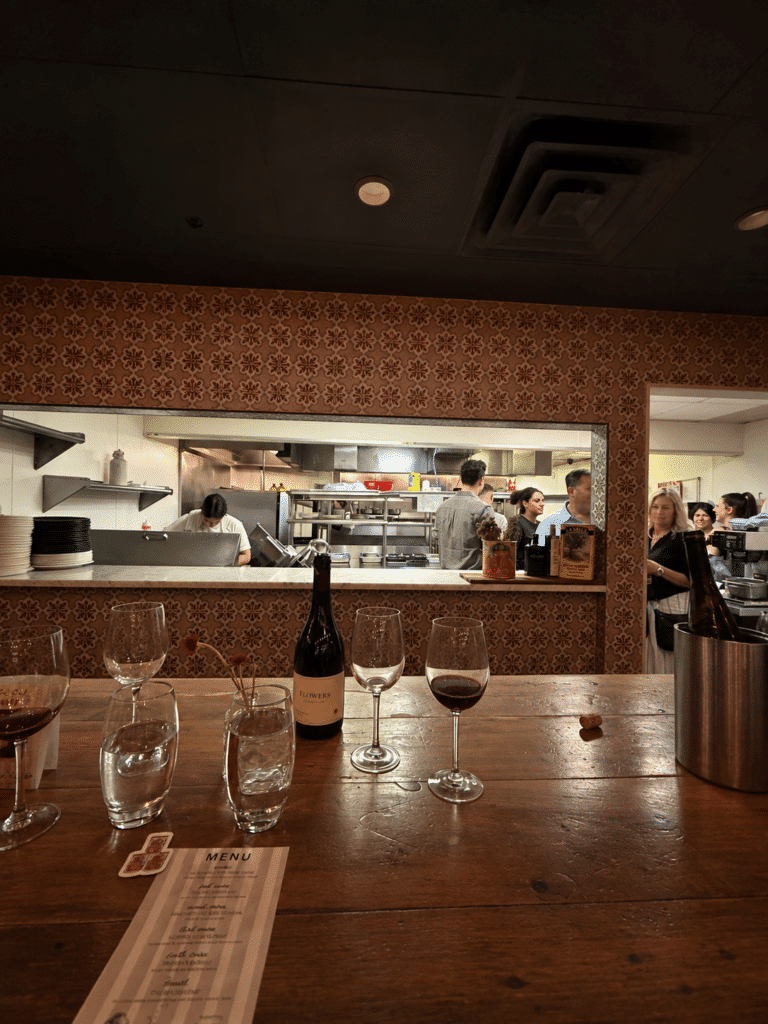 Viaggio x La Valle Dinner Review: Viaggio’s busy open kitchen viewed from the counter, with wine glasses, a Flowers wine bottle and the dinner menu in the foreground.