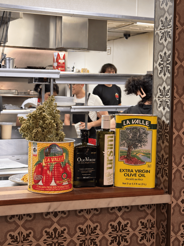 La Valle tomato and olive oil tins beside olive oil bottles, with dried herbs in the tomato tin and kitchen staff behind.