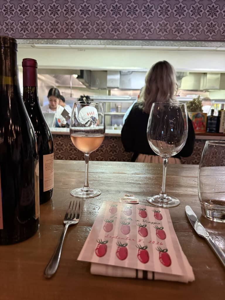 Viaggio x La Valle A glass of rosé, wine bottles and the tomato-illustrated menu at Viaggio’s counter, with the open kitchen behind.