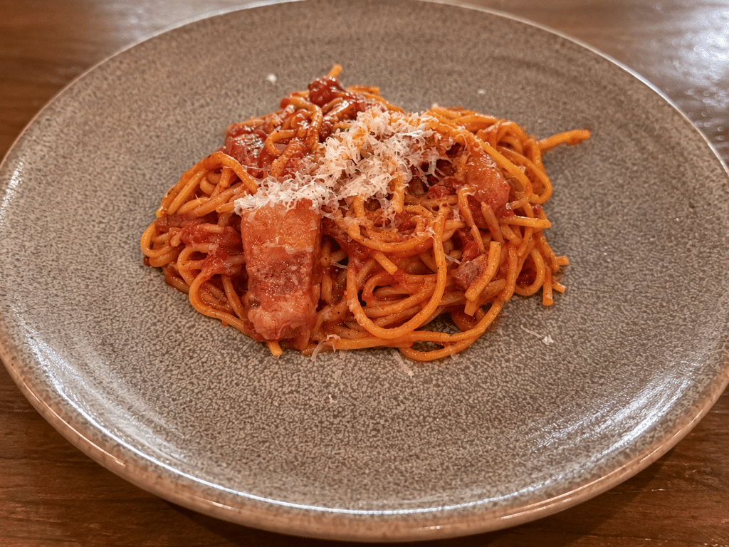 Spaghetti in tomato sauce with guanciale and grated pecorino nero on a textured ceramic plate.