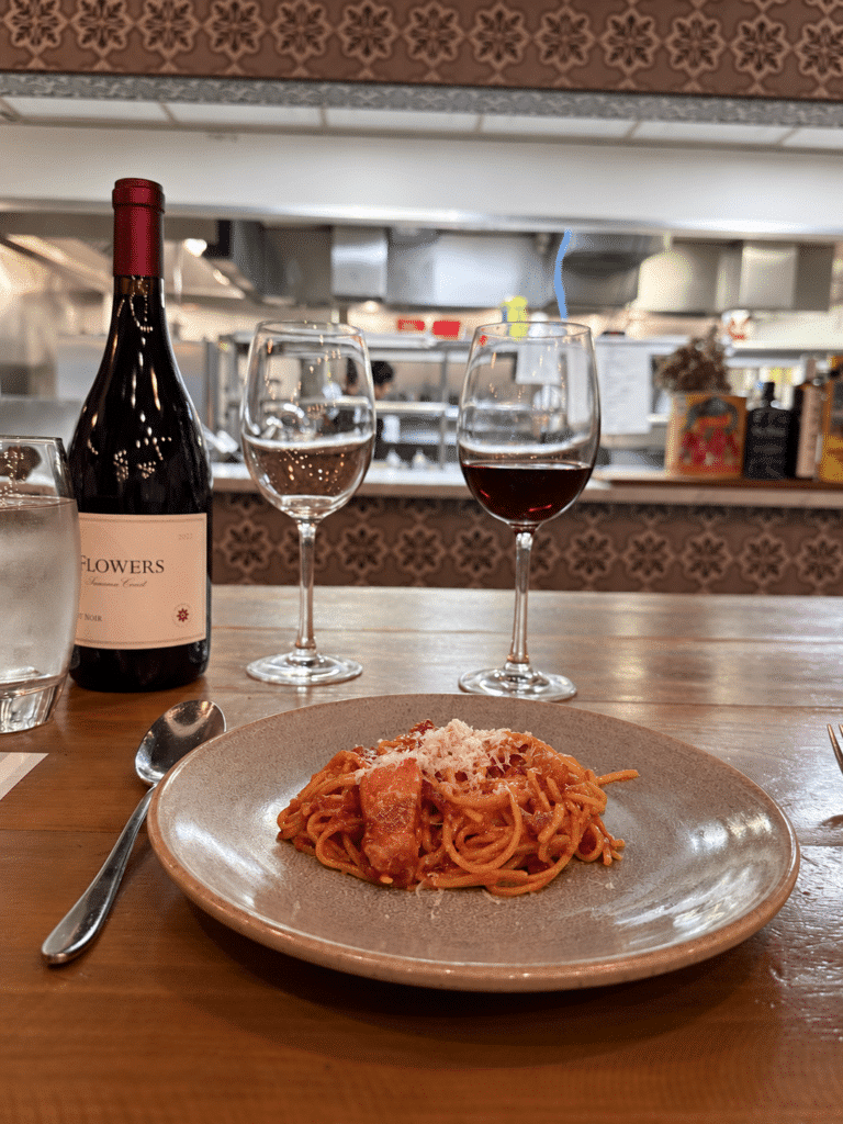 Spaghetti all’Amatriciana beside a Flowers wine bottle and a glass of red wine, overlooking Viaggio’s open kitchen.