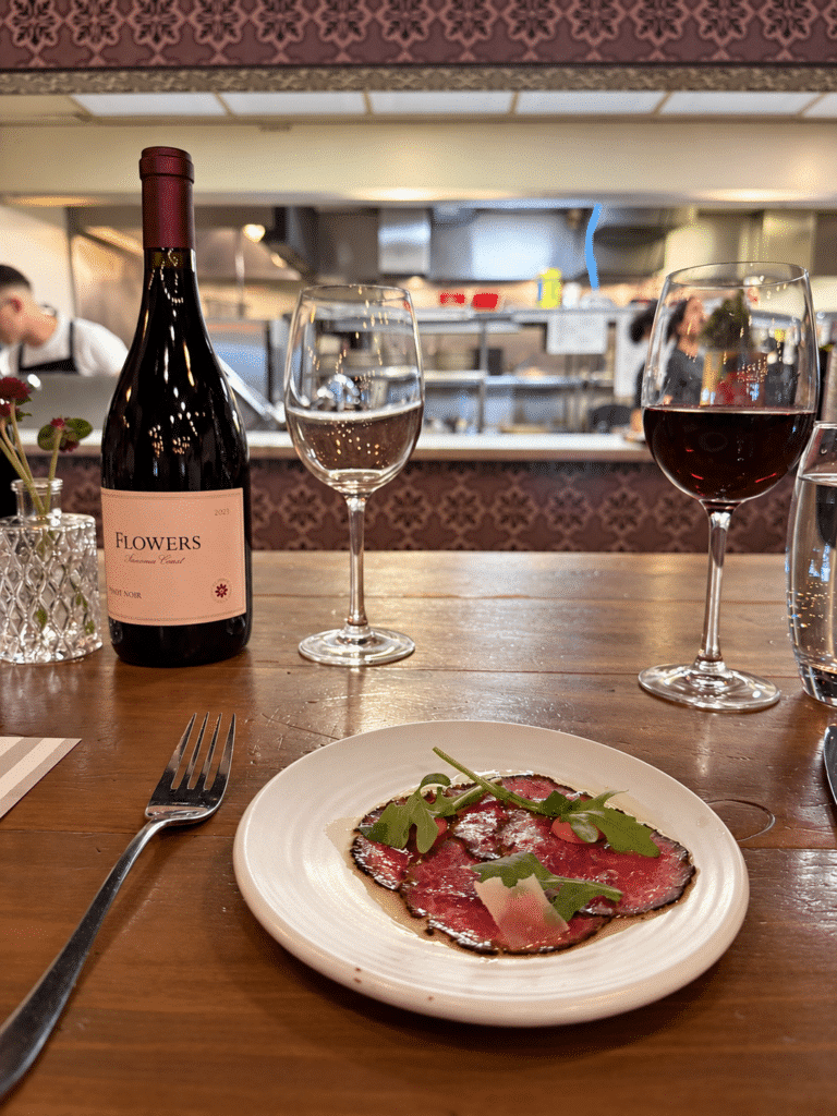 Viaggio x La Valle Dinner Wagyu carpaccio beside a Flowers Pinot Noir bottle and a glass of red wine, with Viaggio’s open kitchen behind.