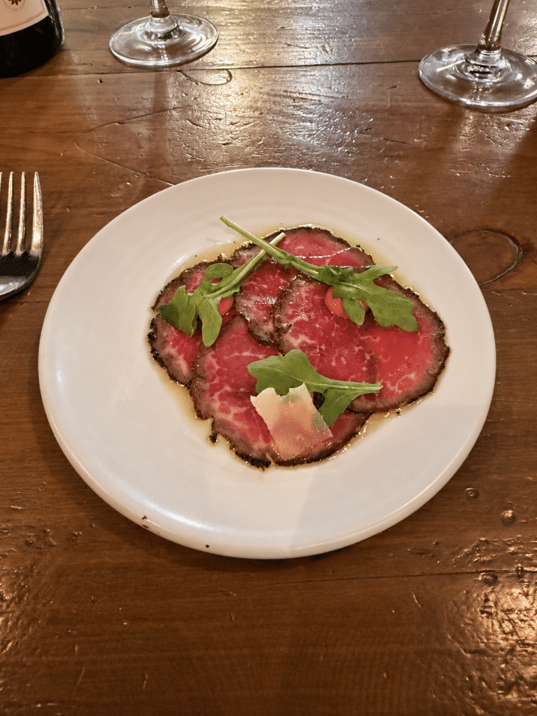 Viaggio x La Valle DInner Thinly sliced Wagyu carpaccio topped with arugula, tomato aioli and shaved Grana Padano on a white plate.