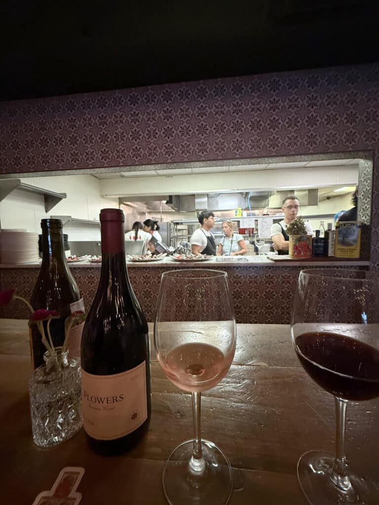 Viaggio x La Valle Dinner Review: A Flowers wine bottle and glasses of rosé and red wine, with staff preparing amuse-bouche portions in Viaggio’s open kitchen behind.