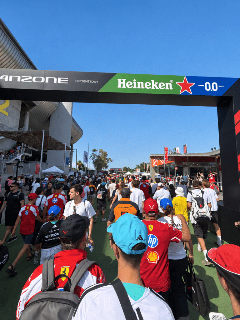 A crowd in Ferrari, McLaren and other team clothing walking beneath the Barcelona fan-zone entrance.
