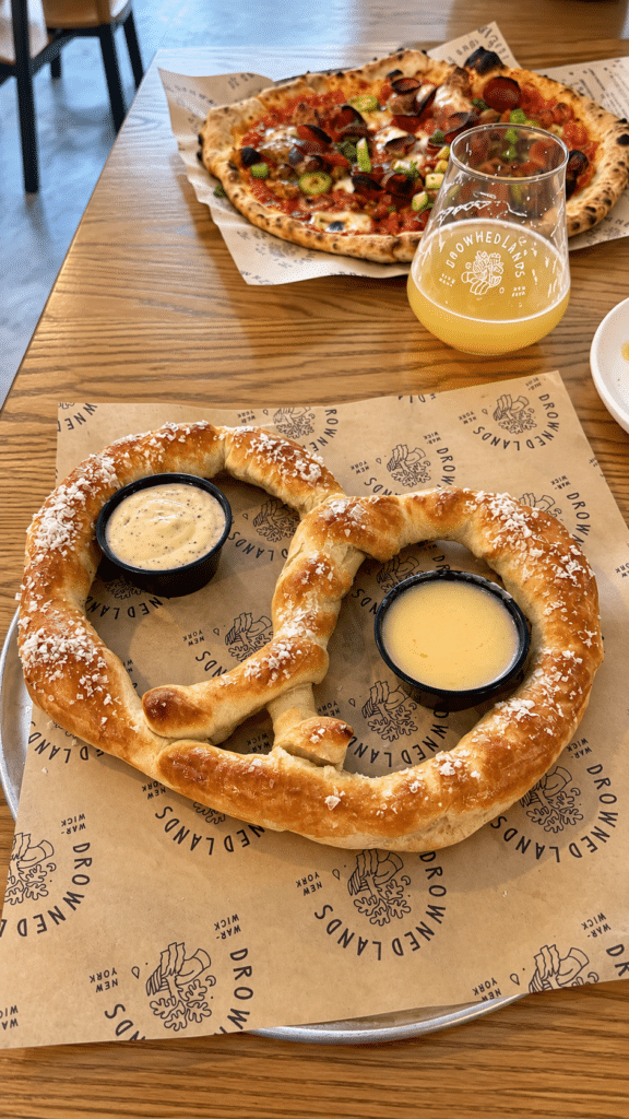 Sourdough pretzel with flaky salt, beer cheese and dijonnaise, with pizza and beer in the background.
