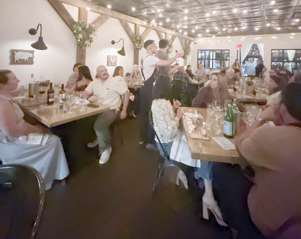 A wide view of guests filling Viaggio's dining room as two speakers address the tables under string lights.