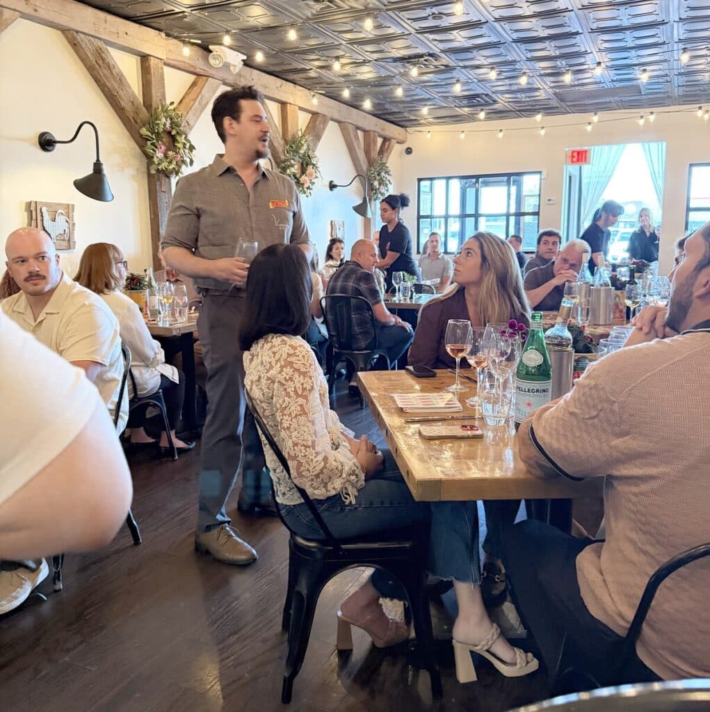 A speaker holding a glass addresses guests seated around wooden tables beneath Viaggio's string lights.