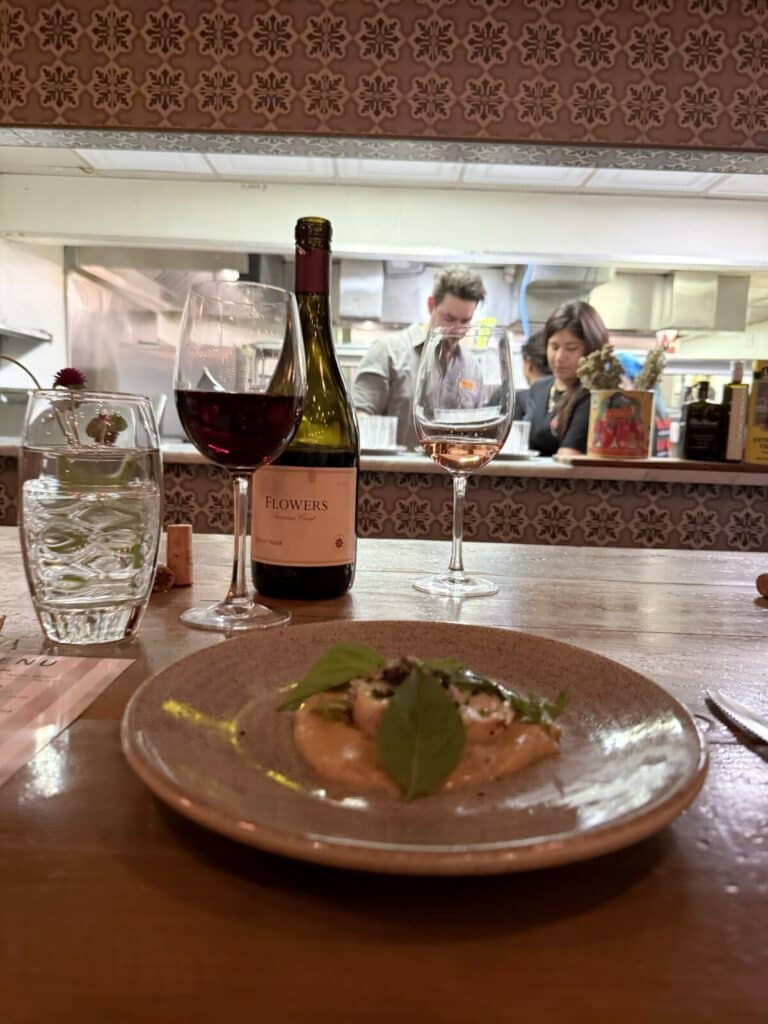 Hiramasa Mosaic, a Flowers wine bottle, and wine glasses at Viaggio's counter, with people in the kitchen behind.