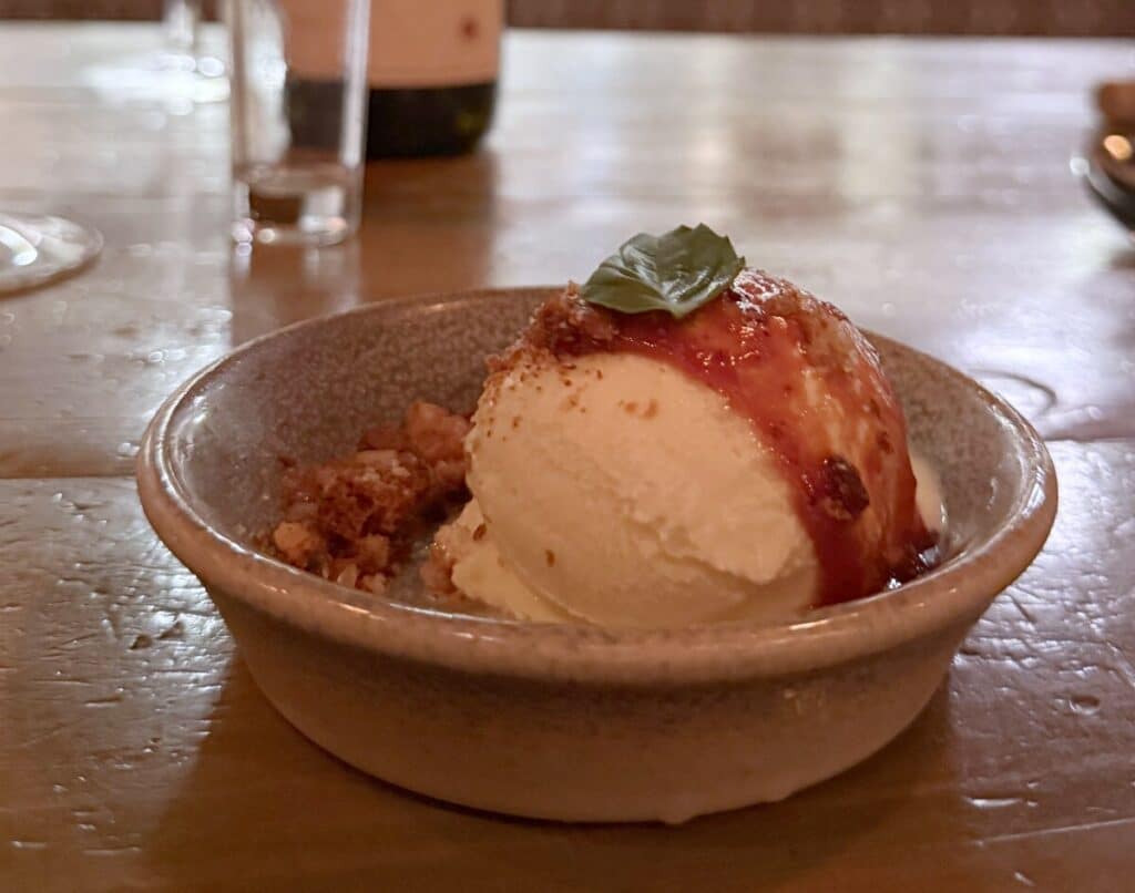 Side view of gelato with tomato jam, basil and focaccia crumble in a shallow bowl on Viaggio's wooden counter.