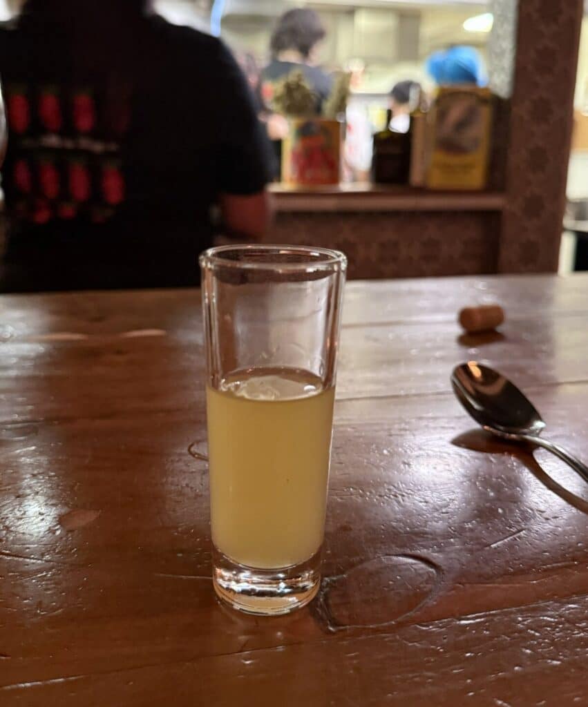 A tall glass of pale yellow limoncello on a wooden counter, with Viaggio's open kitchen softly blurred behind it.