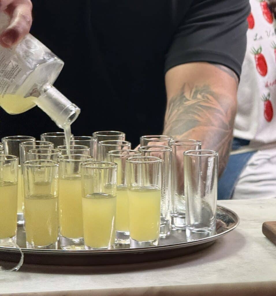 Limoncello being poured from a bottle into tall shot glasses arranged on a silver tray at Viaggio.