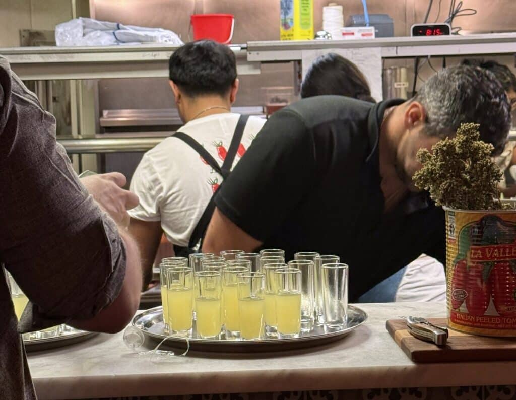 A tray of limoncello glasses on Viaggio's kitchen counter beside a La Valle tomato can, with staff behind the counter.