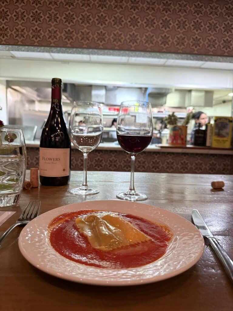 Raviolo Doppio alla Norma beside a Flowers wine bottle and glasses, with Viaggio's open kitchen behind.
