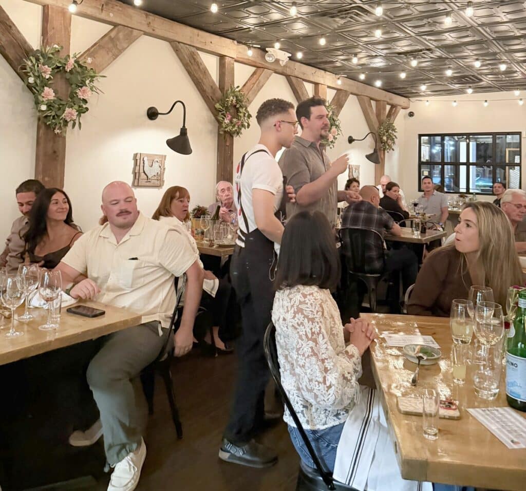 Two speakers standing between tables and addressing seated guests in Viaggio's dining room.
