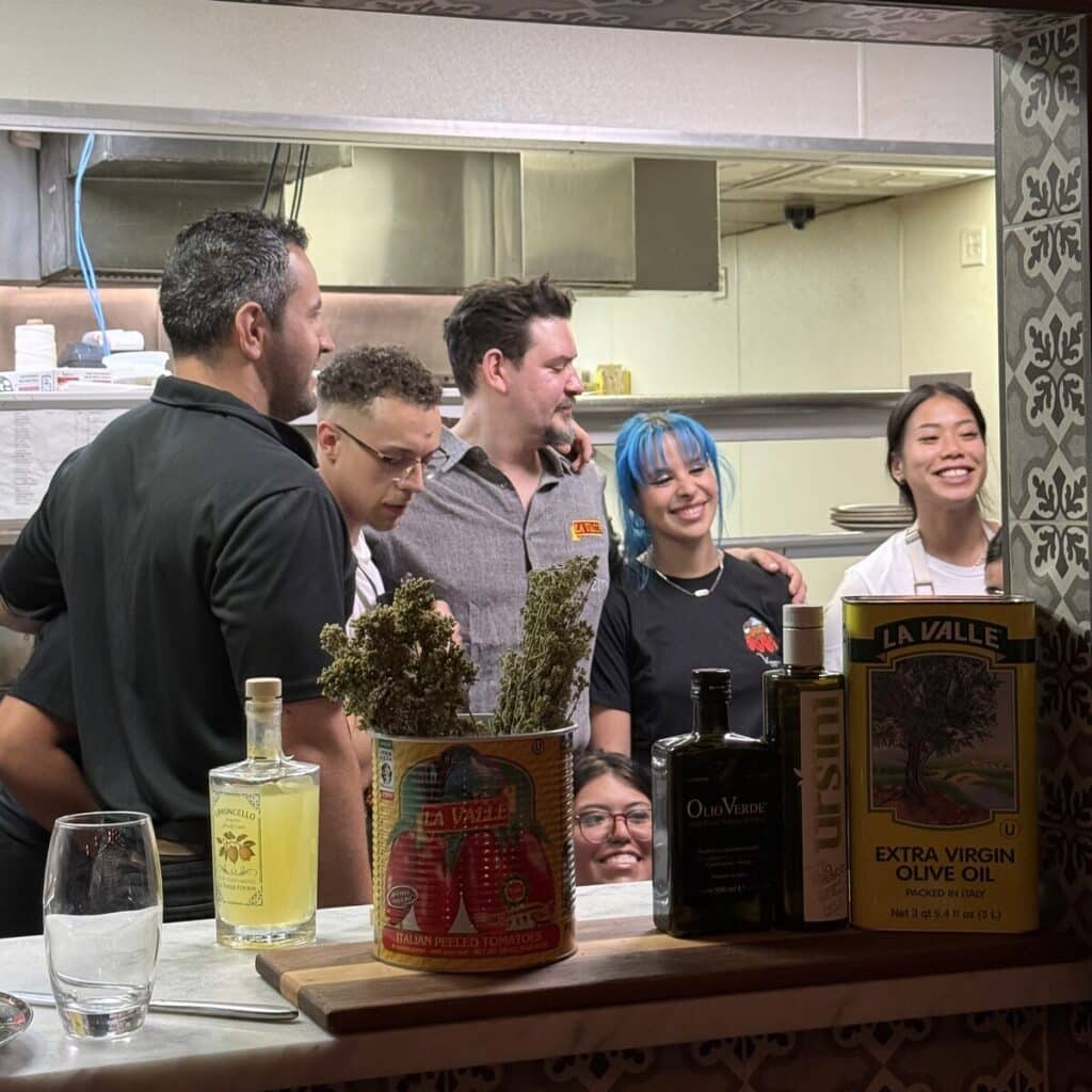 Chef Rabbit, Sam and members of the Viaggio staff posing behind La Valle tomato and olive oil products at the kitchen counter.