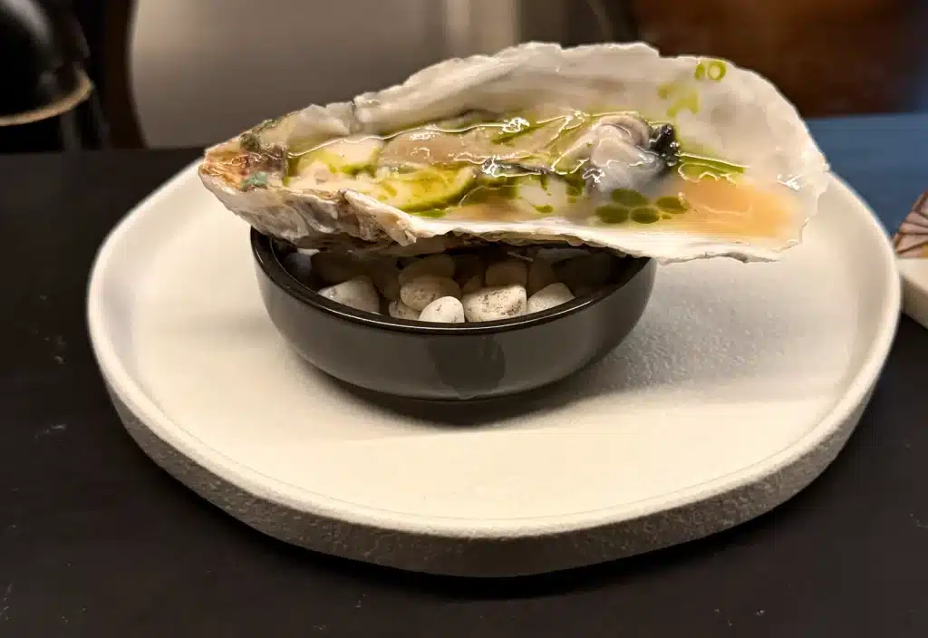 Sushi by Sea  Review Oyster in its shell with green oil droplets, resting above a small black bowl.