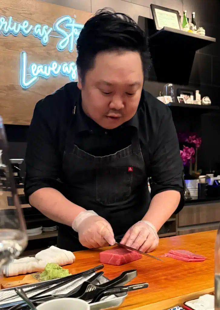 A chef slices a red piece of fish on the wooden counter, with the neon motto behind him.