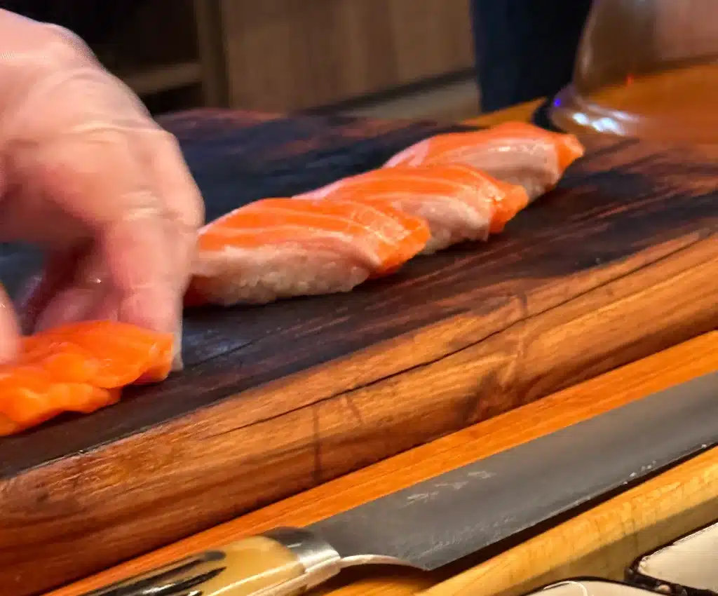 A gloved hand arranges a row of orange salmon nigiri on a wooden board.